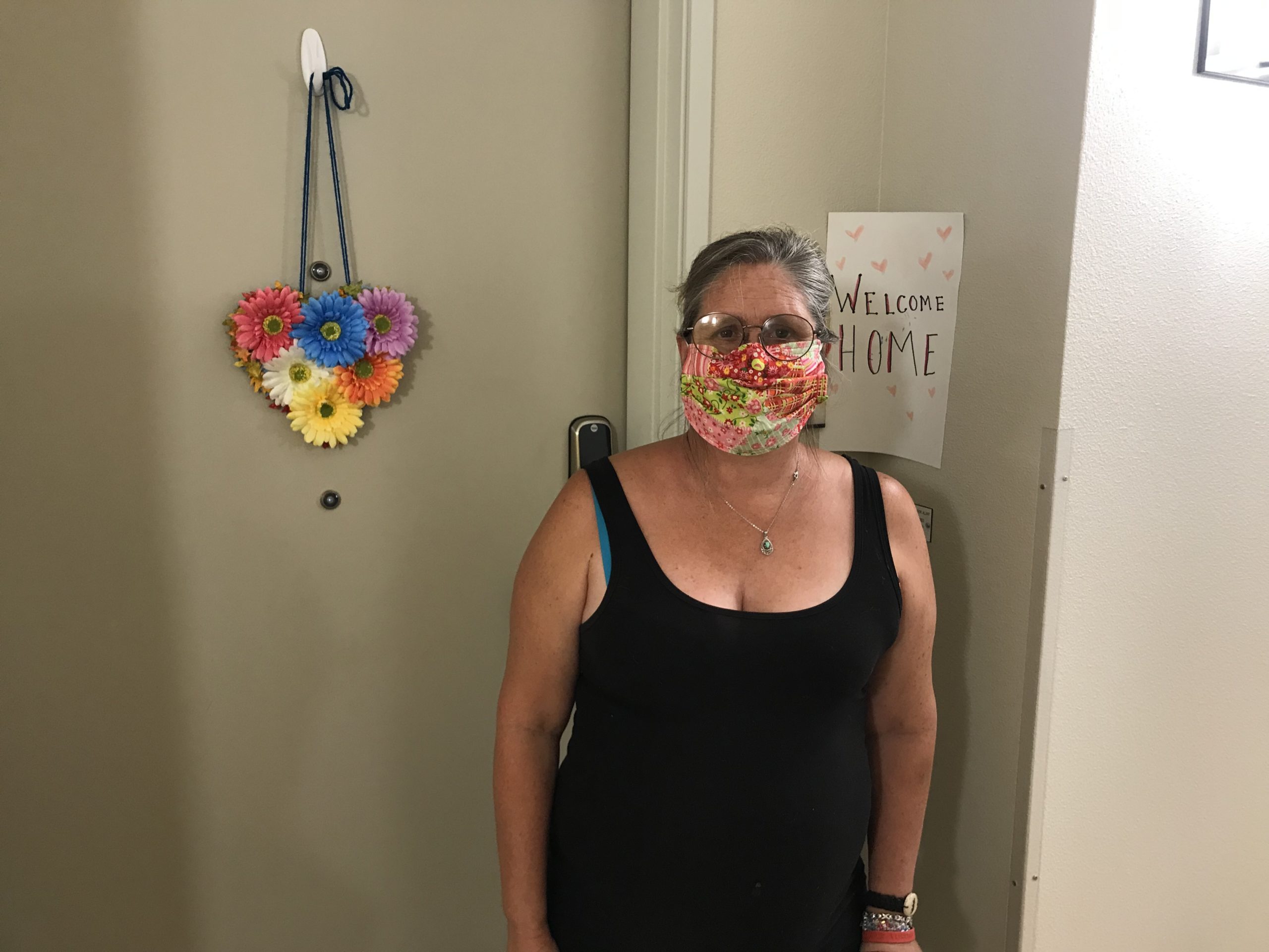 Della with mask on standing in front of her apartment door. "Welcome Home" sign in background.