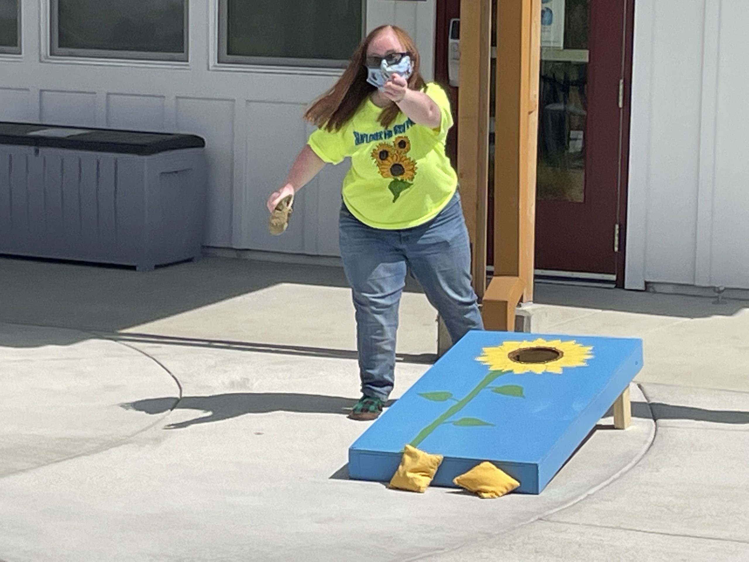 Lauren playing corn hole. Throwing a corn hole bag, standing next to a sunflower-themed corn hole.