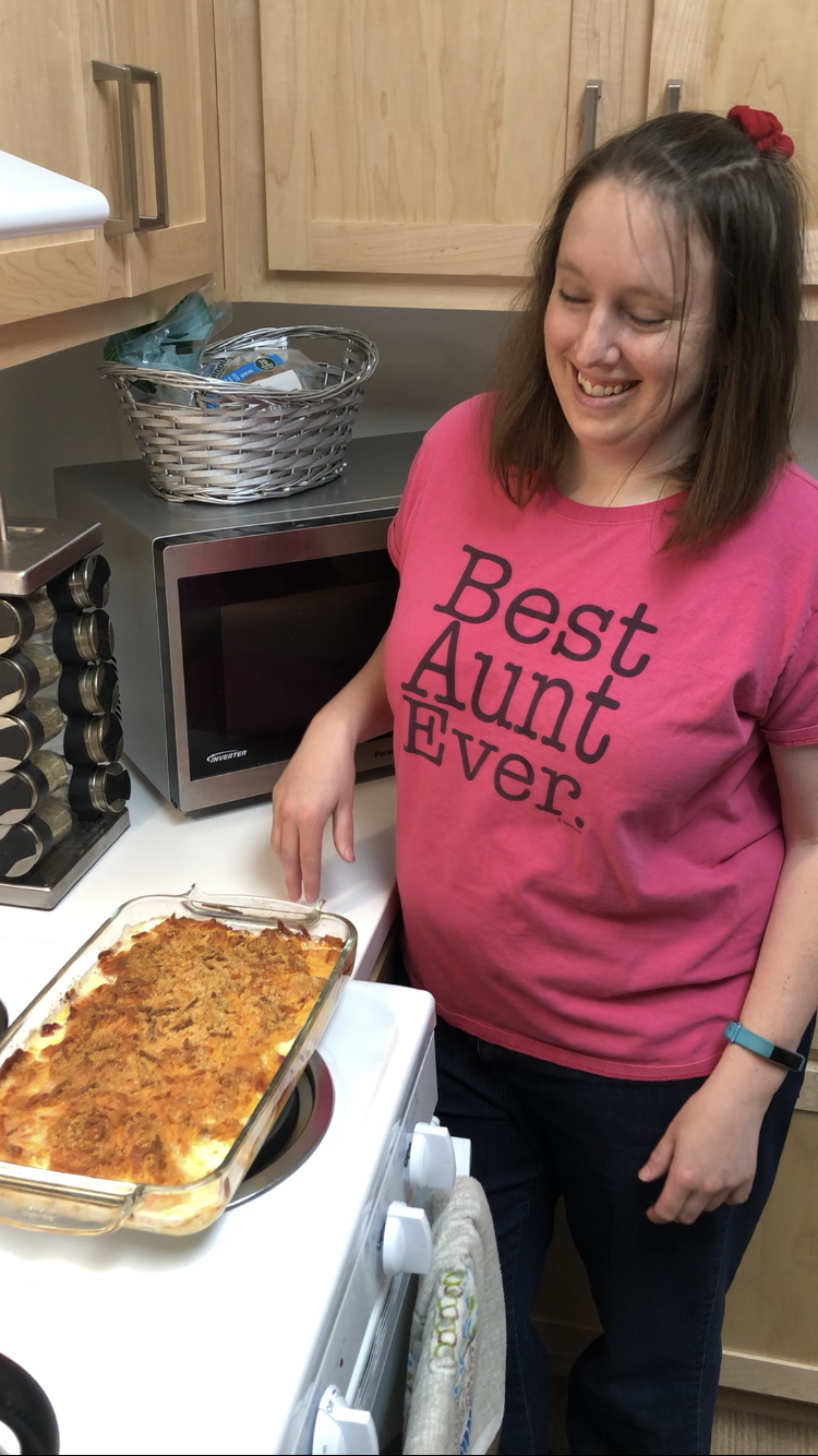 Jennifer in the kitchen standing next to the stove with a casserole she made, wearing  t-shirt that says, "Best Aunt Ever."